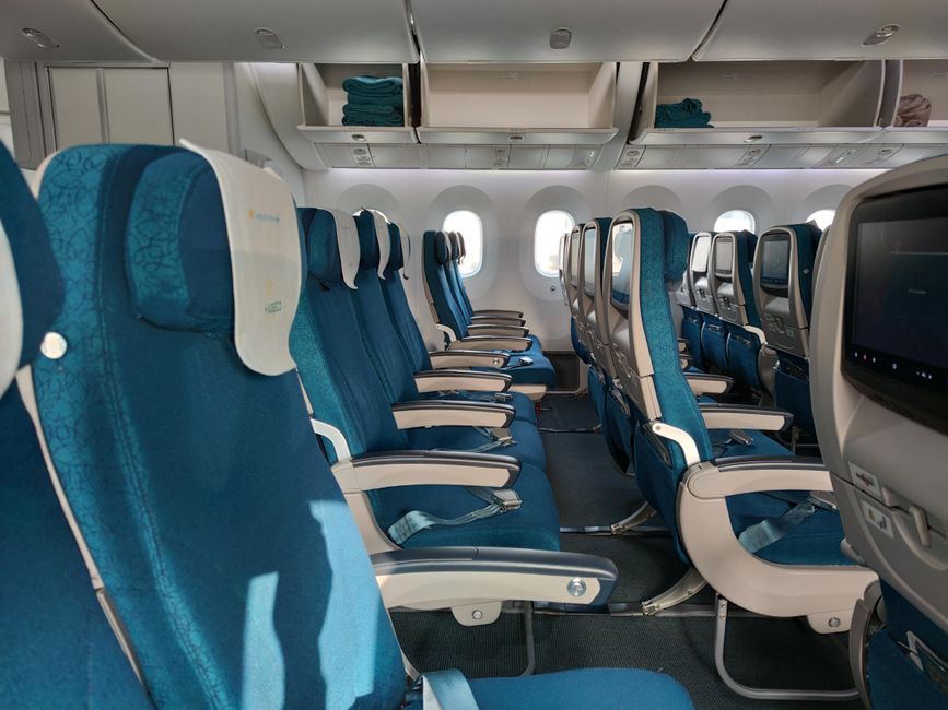 A row of empty blue airplane seats in an economy class cabin on a sunny day