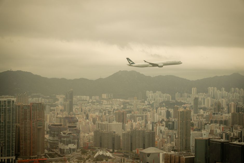 Airplane soars above the bustling skyline of Hong Kong, with mountains in the background.